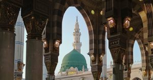 Mescid-i Nebevi'nin süslemeli kemerleri ve sütunları arasından görünen, Peygamber Efendimiz'in kabrinin bulunduğu Yeşil Kubbe (Kubbe-i Hadra) ve Medine silüeti.