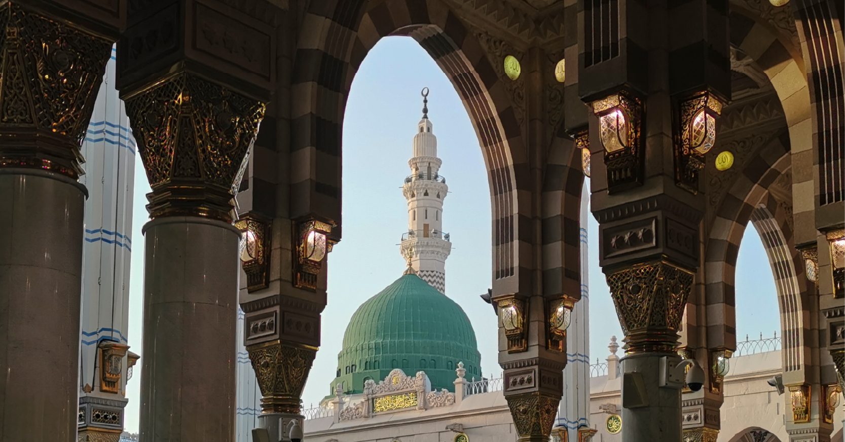 Mescid-i Nebevi'nin süslemeli kemerleri ve sütunları arasından görünen, Peygamber Efendimiz'in kabrinin bulunduğu Yeşil Kubbe (Kubbe-i Hadra) ve Medine silüeti.