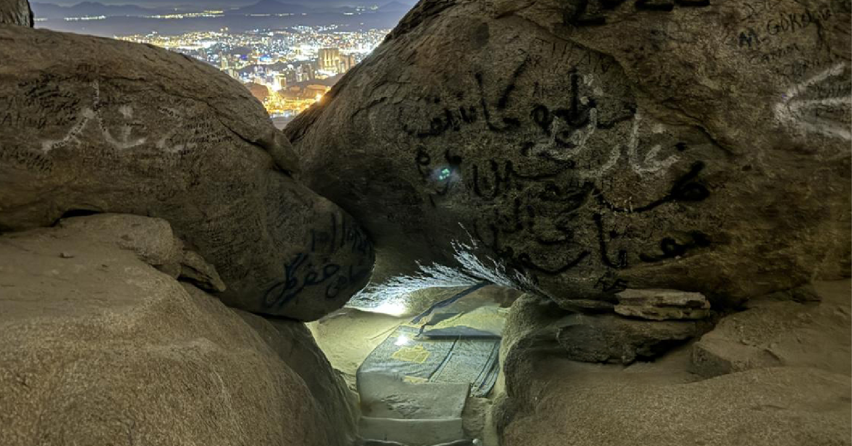 Gece vakti Sevr Mağarası'nın içinden kayaların arasından görünen ışıklı Mekke şehir manzarası ve yerde serili bir seccade.